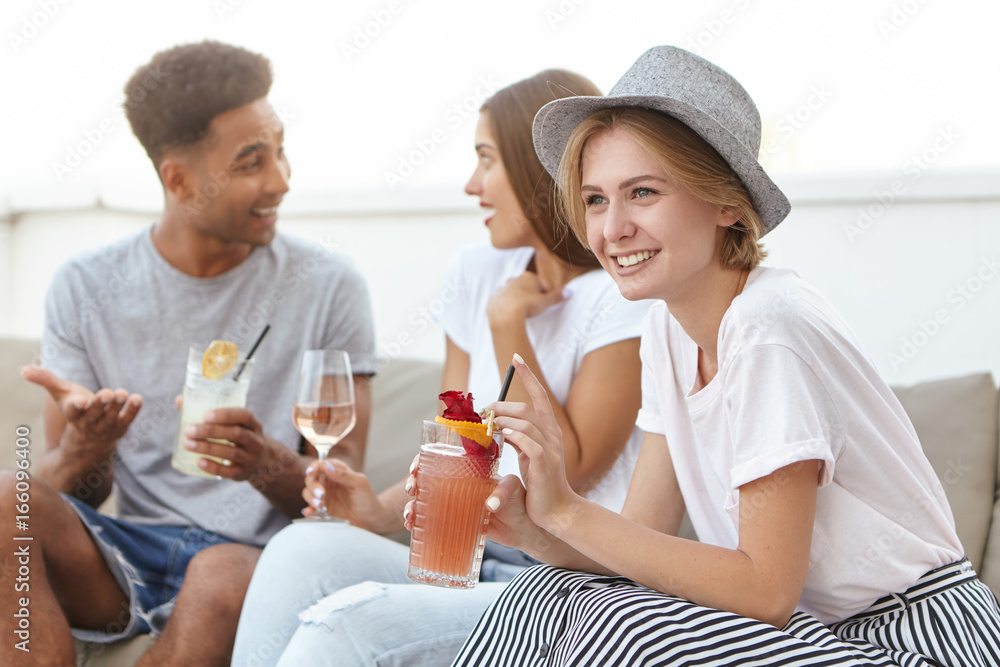 Group of three friends having birthday party sitting at cozy sofa having conversation with each other, drinking alcoholic drinks, having good mood. Happy youngsters celebrating something together Group of three friends having birthday party sitting at cozy sofa having conversation with each other, drinking alcoholic drinks, having good mood. Happy youngsters celebrating something together