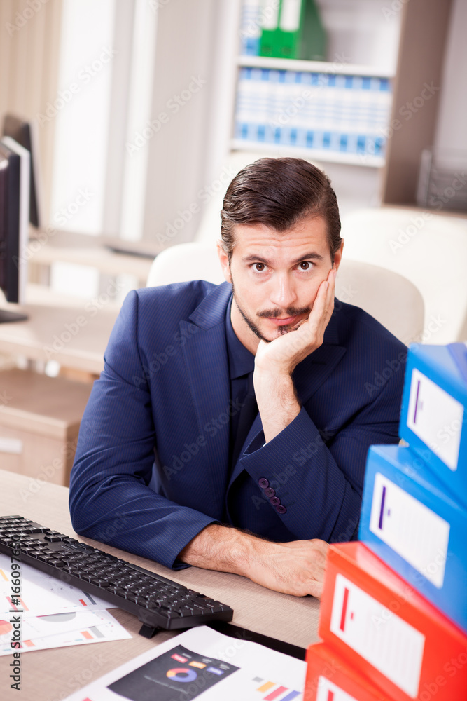 Naklejka premium Young businessman depressed from the workload at the office. His desk is full with a pile of folders and papers with finance charts are on the table.