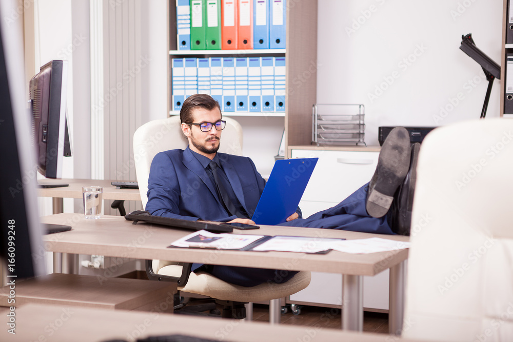 Businessman in office with his feet on the table looking in a blue folder. Image of realaxing corporate worker but still working. Working break Businessman in office with his feet on the table looking in a blue folder. Image of realaxing corporate worker but still working. Working break