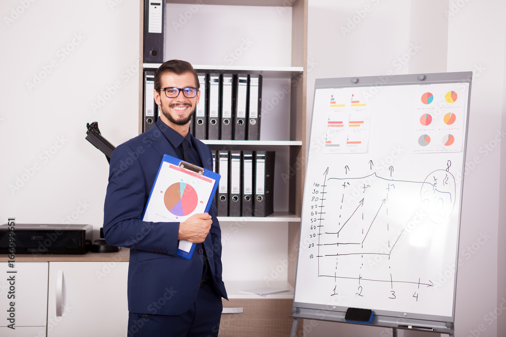 Happy businessman with a folder with charts in hands next to a flip-chart. Image of corporate worker analyzing financial data Happy businessman with a folder with charts in hands next to a flip-chart. Image of corporate worker analyzing financial data
