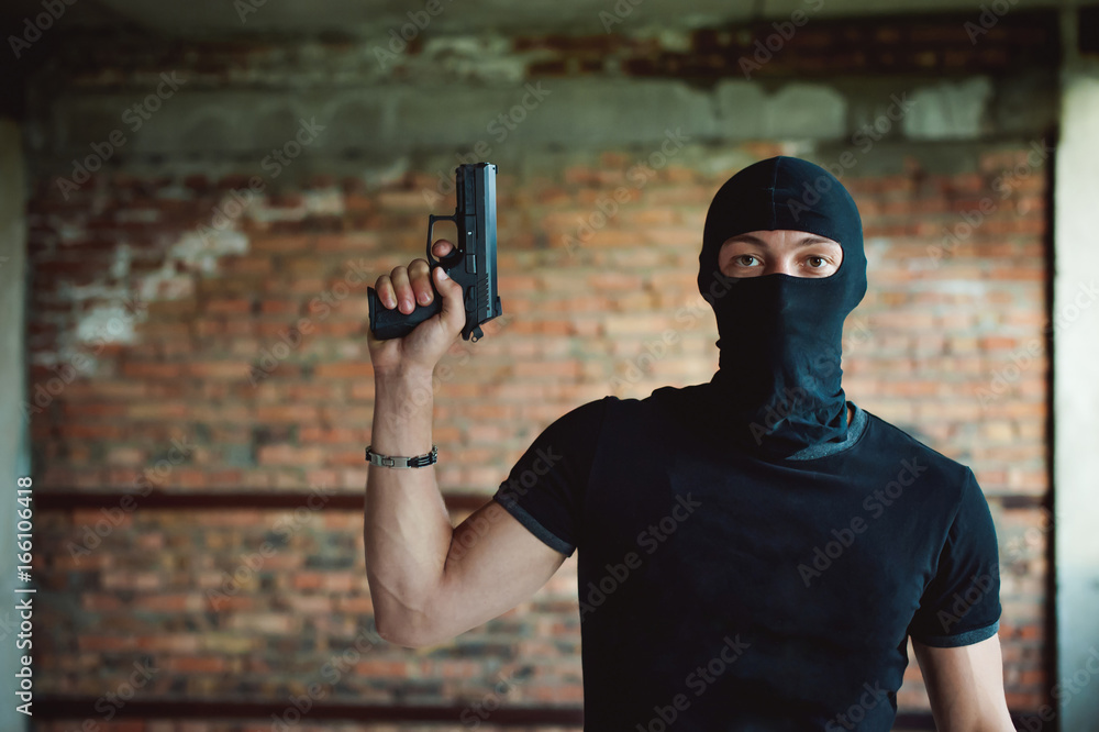 Athletic man in a balaclava, ski mask on his head, holds a pistol in his  hand, lifting up a barrel, a dangerous gangster foto de Stock | Adobe Stock