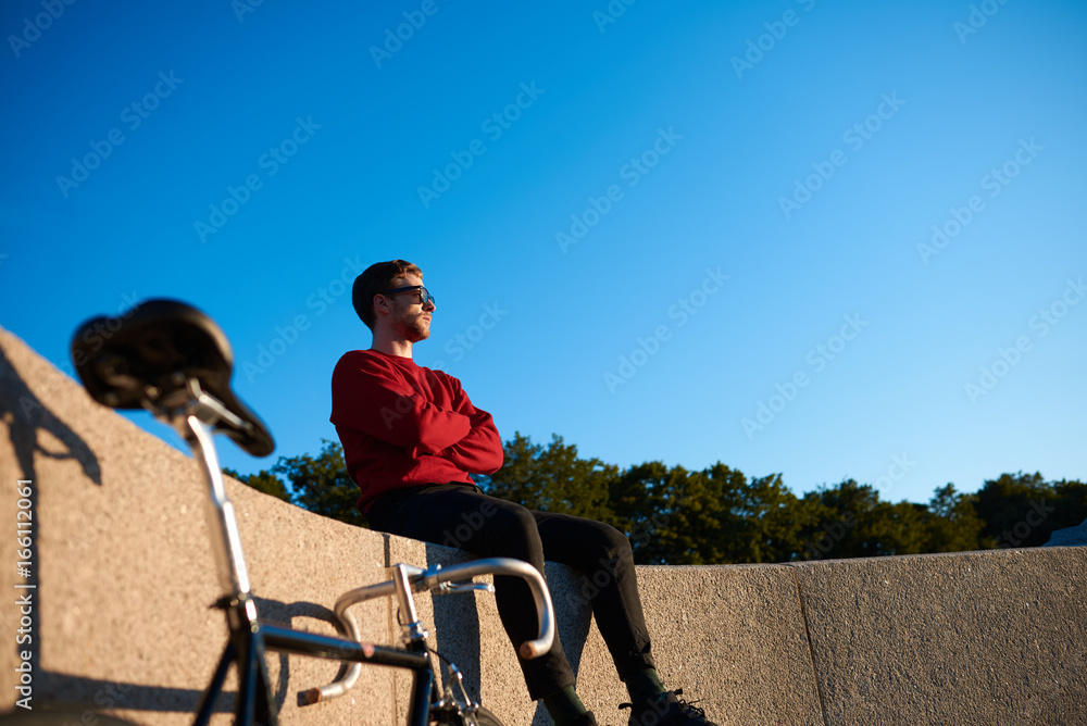 Outdoor shot of serious confident young hipster wearing shades sitting on concrete fencing with his fixed gear bike next to him, looking into distance, contemplating beautiful nature around him