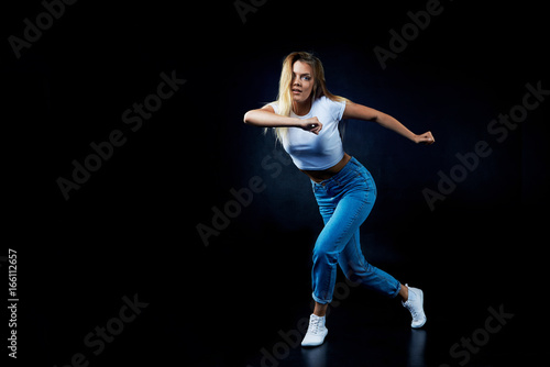 Full lenght portrait of flexible slim gorgeous female with blonde loose hair dancing hip-hop in dark studio with copy space for your text or promtional content. Dance, performance and aerobics