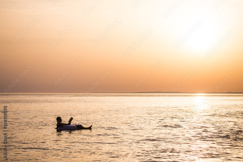 Young man in relax in the ocean water on inflatable ring on sunset read a book.