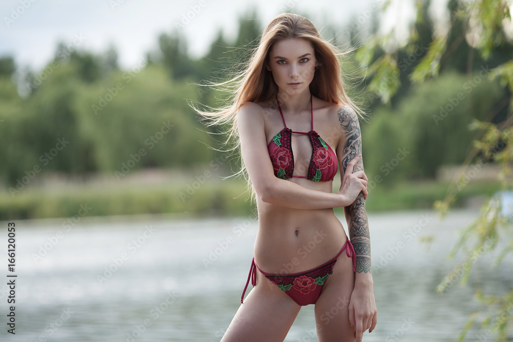 Sensual tattooed young woman posing in lingerie, looking at camera. Sexy girl in lingerie on sunset in the woods in nature.