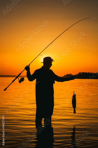 A fisherman with a fishing rod in his hand and a fish caught stands in the water against a beautiful sunset.