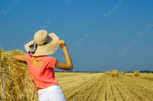 The girl is standing near a straw bale and looking into the distance in the field photo