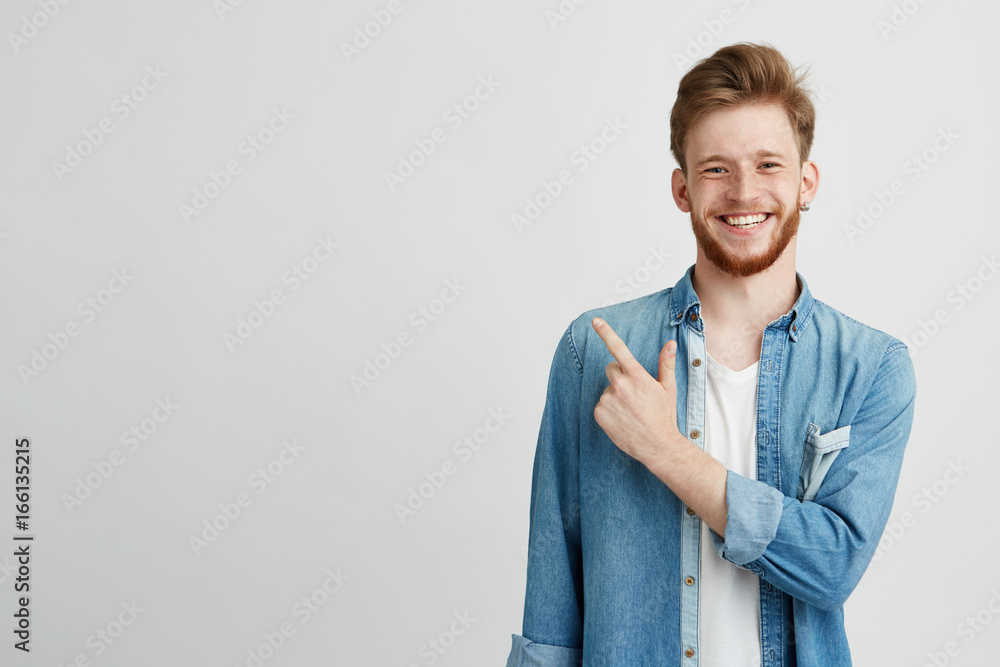 Portrait of cheerful young man smiling looking at camera pointing finger up over white background. - obrazy, fototapety, plakaty 