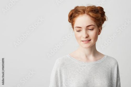 Portrait of shy young pretty redhead girl with buns looking down smiling over white background. 