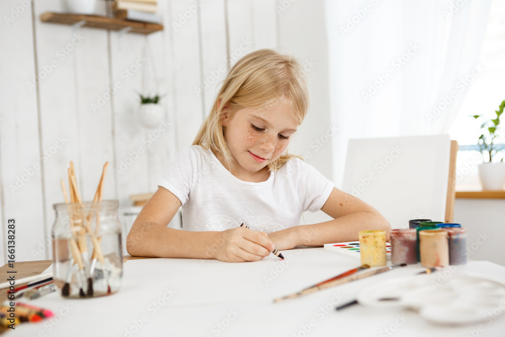 Indoor portrait of cute little blonde haired girl with freckles drawing with crayon color on the sheet of paper. Blonde child wearing white T-shirt sitting at the table drawing a picture. Indoor portrait of cute little blonde haired girl with freckles drawing with crayon color on the sheet of paper. Blonde child wearing white T-shirt sitting at the table drawing a picture.