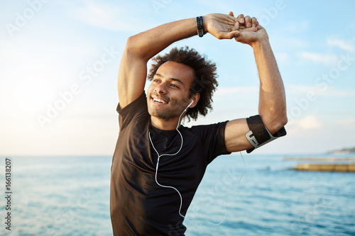 Portrait of a smiling afro-american sports man stretching his muscular arms before workout by the sea, using music app on his smartphone. Dark-skinned athlete warming up before running.