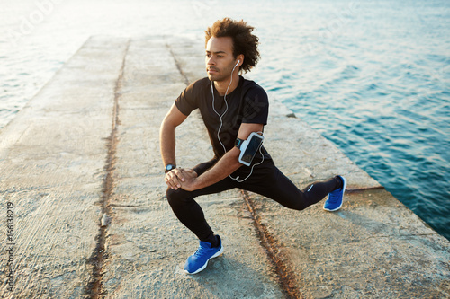 Young male dark-skinned runner with beautiful fit body warming-up his muscles before strength training cardio workout. Man athlete in sportswear stretching legs with lunge hamstring stretch exercise