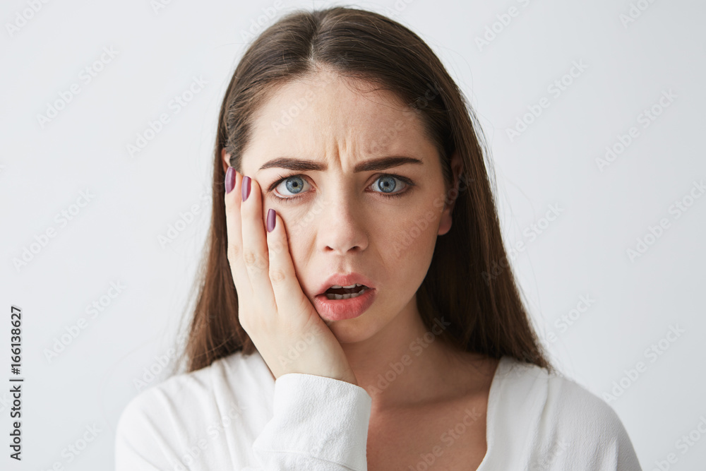 Close up photo of displeased frightened girl looking at camera touching face frowning over white background. Close up photo of displeased frightened girl looking at camera touching face frowning over white background.