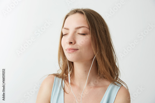 Tender young pretty girl smiling with closed eyes listening to streaming music in headphones over white background.