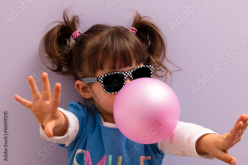 Little girl with sun glasses blowing up pink chewing gum against a light purple background photo