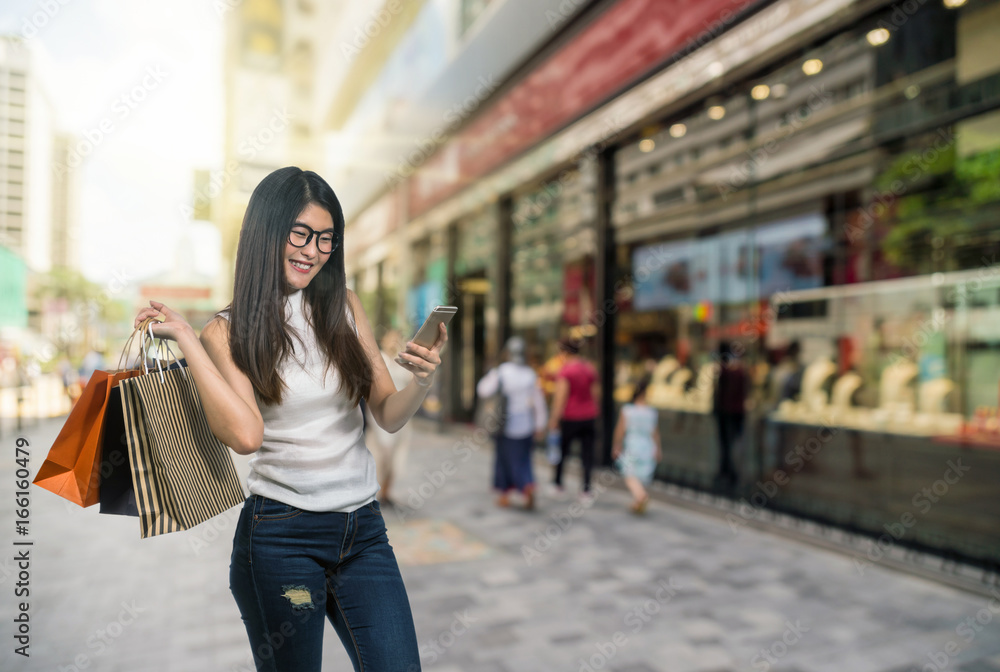 Fototapeta premium Happy young asian woman shopping in happy feeling and holding the product paper bag over Abstract blurred photo of shopping store in outdoor store mall with people walking streer, shopping concept