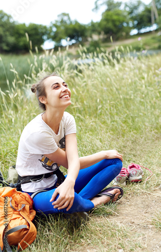 young smiling woman climber in climbing harness seats on the grass, having rest and looking up