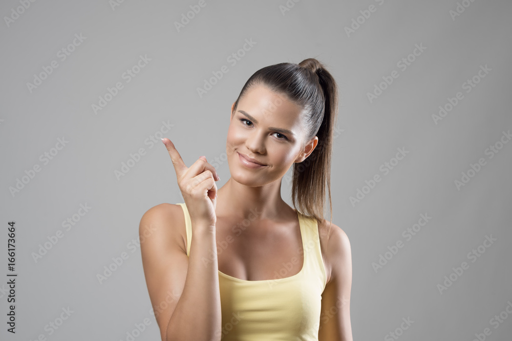 Smiling young sporty woman in yellow tank top scolding finger and looking at camera over gray studio background. Smiling young sporty woman in yellow tank top scolding finger and looking at camera over gray studio background.