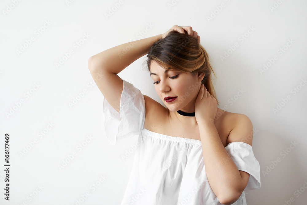 Beauty and makeup concept. Beautiful tender young European woman model wearing white top with open shoulders and black choker, looking down and adjusting her hairstyle while posing in studio Beauty and makeup concept. Beautiful tender young European woman model wearing white top with open shoulders and black choker, looking down and adjusting her hairstyle while posing in studio
