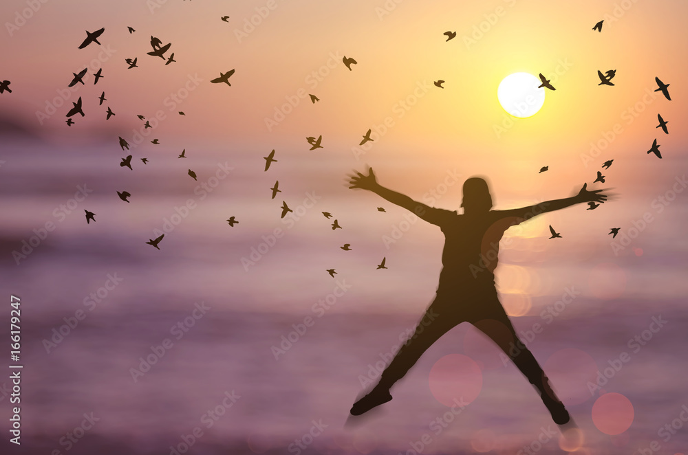 Freedom and feel good concept. Copy space of silhouette happy man jumping on blur tropical sunset beach with birds fly abstract background.