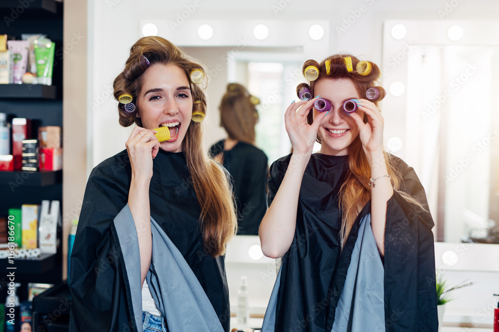Φωτογραφία Two funny young girlfriends in hair curlers wearing capes having fun time together in beauty salon