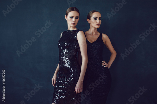 Two beautiful women in black night fashion dress posing on a black background.