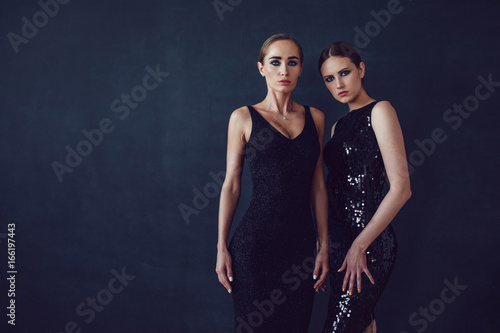 Two beautiful women in black night fashion dress posing on a black background.