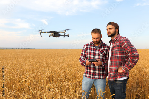 Compact drone hovers in front of two hipster men. Quadcopter flies near farmer and agronomist exploring harvest with innovative technology taking aerial photos and videos from above it wheat field