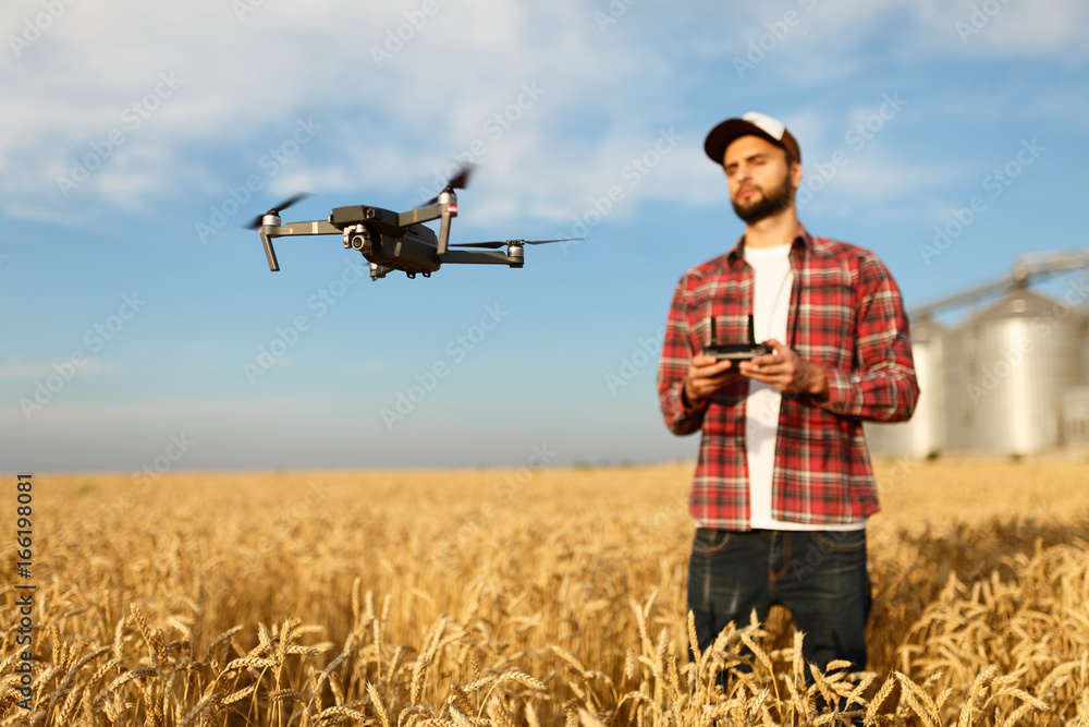 Drone hovers in front of farmer with remote controller in hands near grain elevator. Quadcopter flies near pilot. Agronomist taking aerial photos and videos in a wheat field. Drone hovers in front of farmer with remote controller in hands near grain elevator. Quadcopter flies near pilot. Agronomist taking aerial photos and videos in a wheat field.