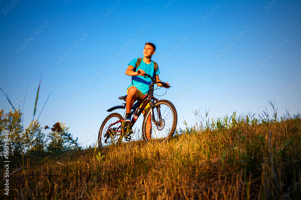 The boy on a bicycle is looking the sunset. The boy is standing on the hillside, behind him a blue sky without clouds. The boy on a bicycle is looking the sunset. The boy is standing on the hillside, behind him a blue sky without clouds.