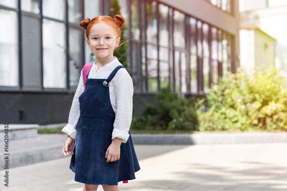 portrait of beautiful small red haired schoolgirl in eyeglasses looking at camera outside school portrait of beautiful small red haired schoolgirl in eyeglasses looking at camera outside school