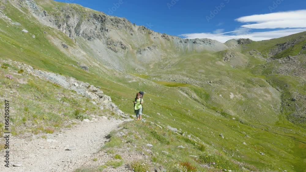 Woman trekking in idyllic mountain landscape on footpath crossing blooming green meadow set amid high altitude rocky mountain range and peaks. Summer adventures on the Italian Alps. Woman trekking in idyllic mountain landscape on footpath crossing blooming green meadow set amid high altitude rocky mountain range and peaks. Summer adventures on the Italian Alps.