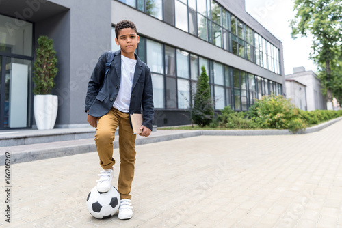 cute african american scoolboy standing with soccer ball and looking at camera near school photo