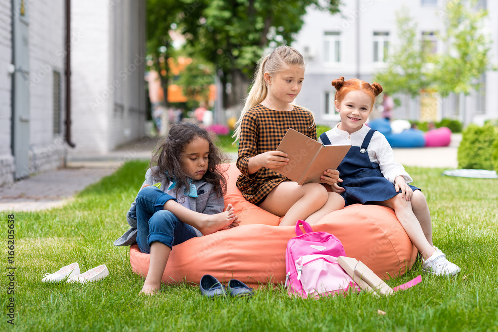 adorable multiethnic schoolgirls sitting on bean bag chair and reading book on schoolyard
