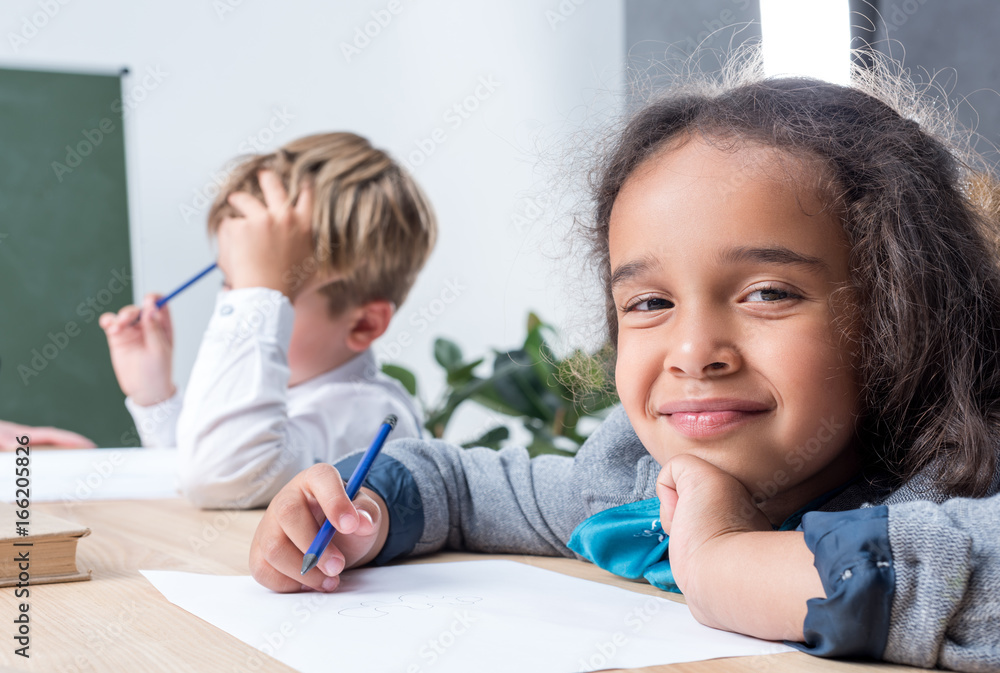 cute multiethnic schoolkids drawing while sitting at desk in class cute multiethnic schoolkids drawing while sitting at desk in class