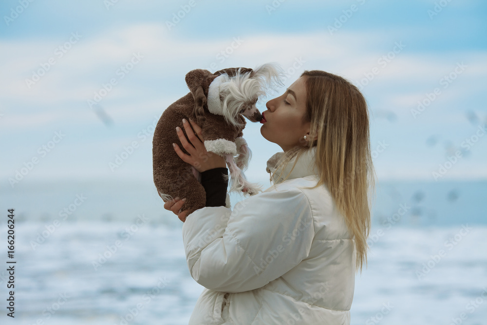 Winter vacation concept. Couple of friends standing on beach with flying seagulls near water. Girl kissing her Chinese crested dog. Hipster style. Happy together. Outdoor shot