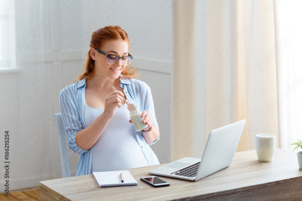 smiling redhead pregnant woman eating yoghurt while sitting at table with laptop, notepad and smartphone at home smiling redhead pregnant woman eating yoghurt while sitting at table with laptop, notepad and smartphone at home
