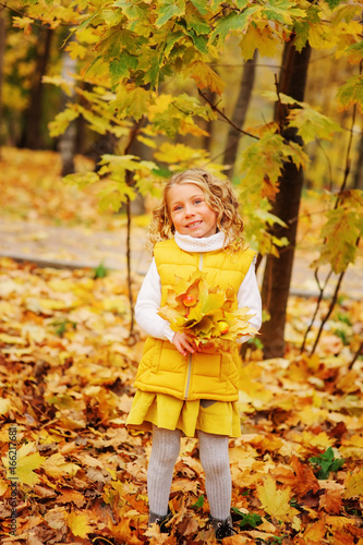 cute toddler girl playing with leaves in autumn park on the walk, wearing fashion yellow outfit