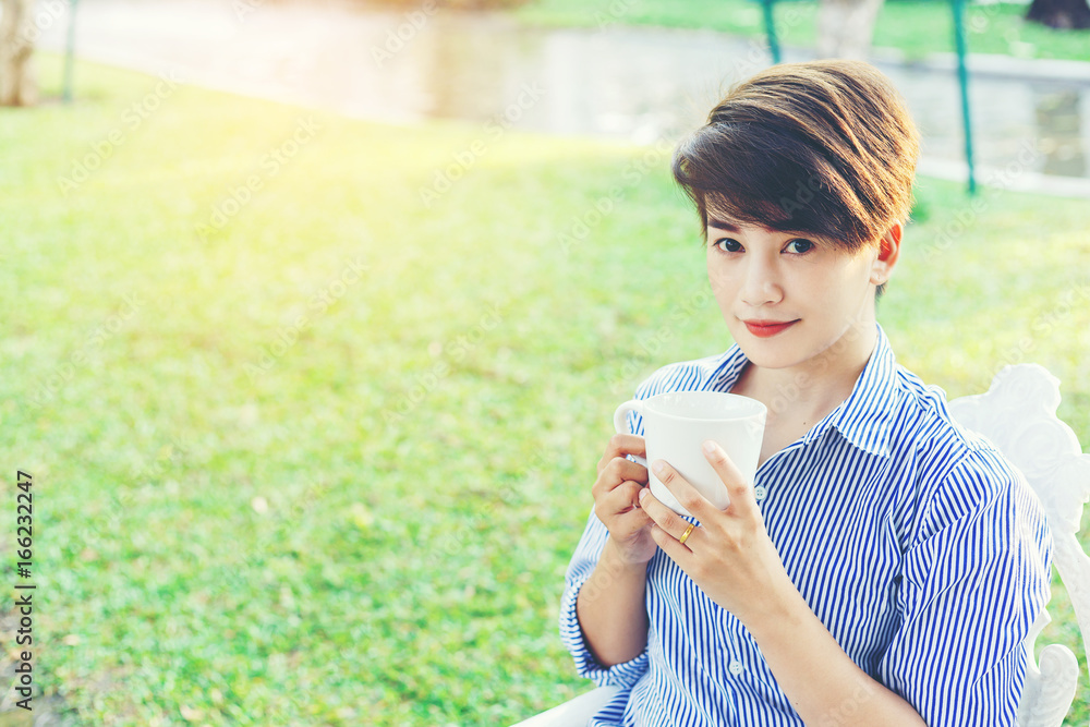 Beautiful short hair woman sitting on a white chair and drinking a favorite beverage in white cup for resting between her outdoor working business in the park. Beautiful short hair woman sitting on a white chair and drinking a favorite beverage in white cup for resting between her outdoor working business in the park.