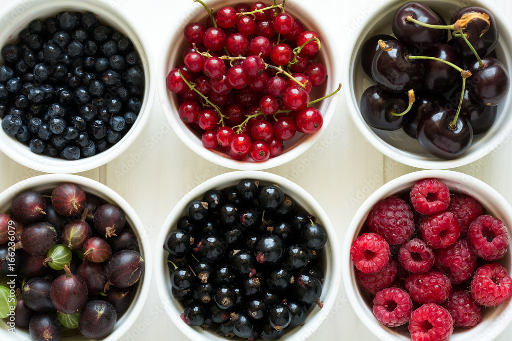 Bowls with fresh ripe gooseberry, red currant, black currant, raspberry, blueberry and cherry on white wooden table Bowls with fresh ripe gooseberry, red currant, black currant, raspberry, blueberry and cherry on white wooden table