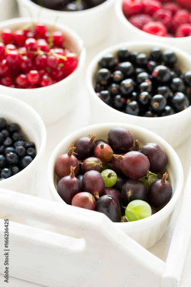Bowls with fresh ripe gooseberry, red currant, black currant, raspberry, blueberry and cherry on white wooden table Bowls with fresh ripe gooseberry, red currant, black currant, raspberry, blueberry and cherry on white wooden table