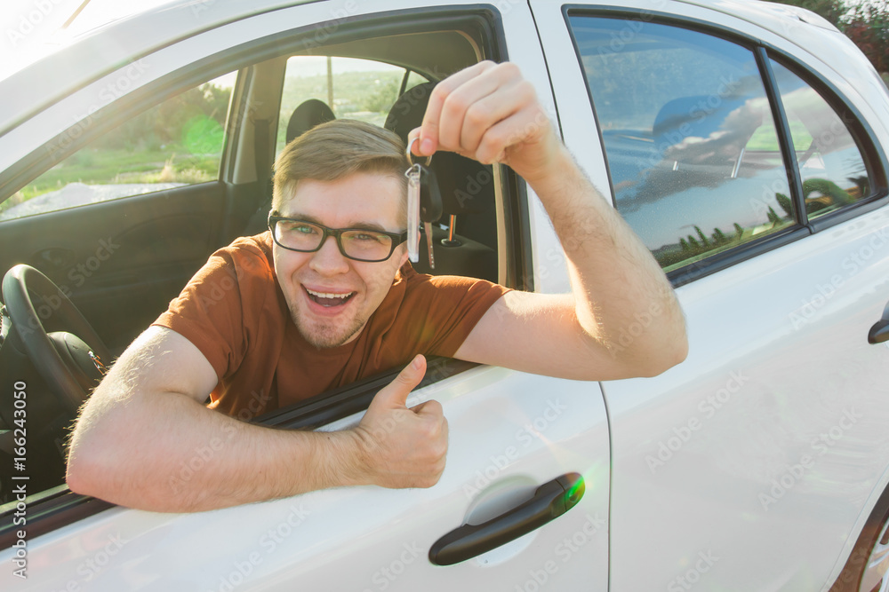 Naklejka premium Young driver showing car keys and thumbs up happy. Man holding car key for new automobile. Rental cars or drivers licence concept with male driving in beautiful nature on road trip.
