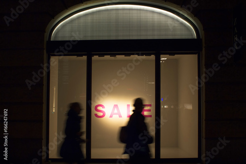 Silhouettes of women walking in blurry motion in front of a fashion store's window in night. Conceptual image of sale season and consumerism, capitalism of modern world.