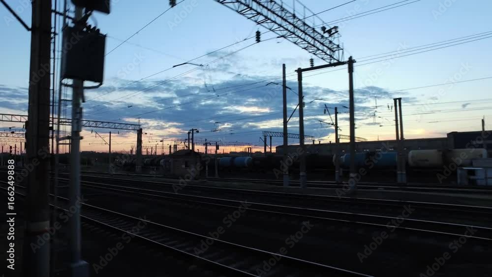 The train is approaching the railway station in the background of a sunset by the side of a freight train standing on the neighboring tracks. The train is approaching the railway station in the background of a sunset by the side of a freight train standing on the neighboring tracks.