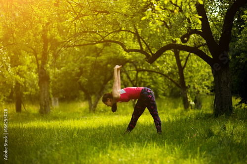 Woman doing Intense Side Stretch Pose practicing yoga. Early morning garden woman doing yoga pose Parsvottanasana. Toned image.