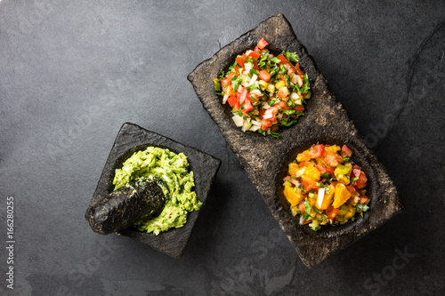 Famous mexican sauces salsas - pico de gallo, avocado guacamole, salsa bandera mexicana in stone mortars on gray slate background