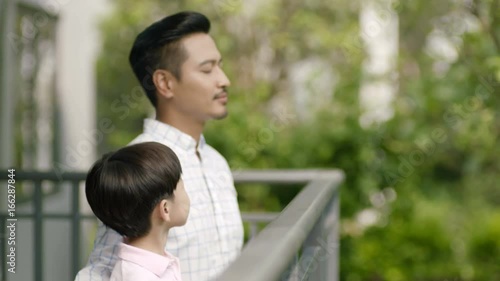 Asian father & son standing on balcony, smiling, looking at each other and taking deep breath