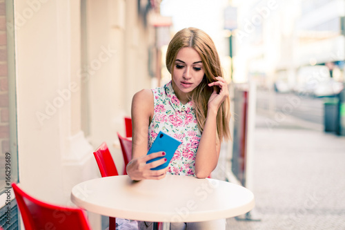 Girl blonde is sitting at an empty table of a street cafe with a smartphone in her hand © luchschenF