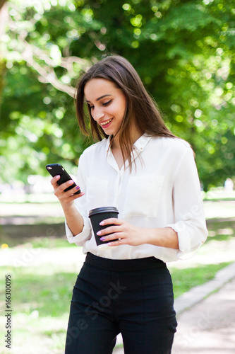 Girl walks with phone in her hand and a cup of coffee in the park