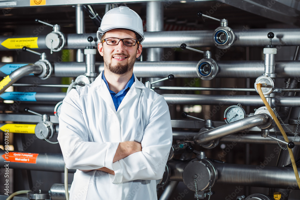 Portrait of a young smiling male worker in a white coat and helmet at a beer factory.